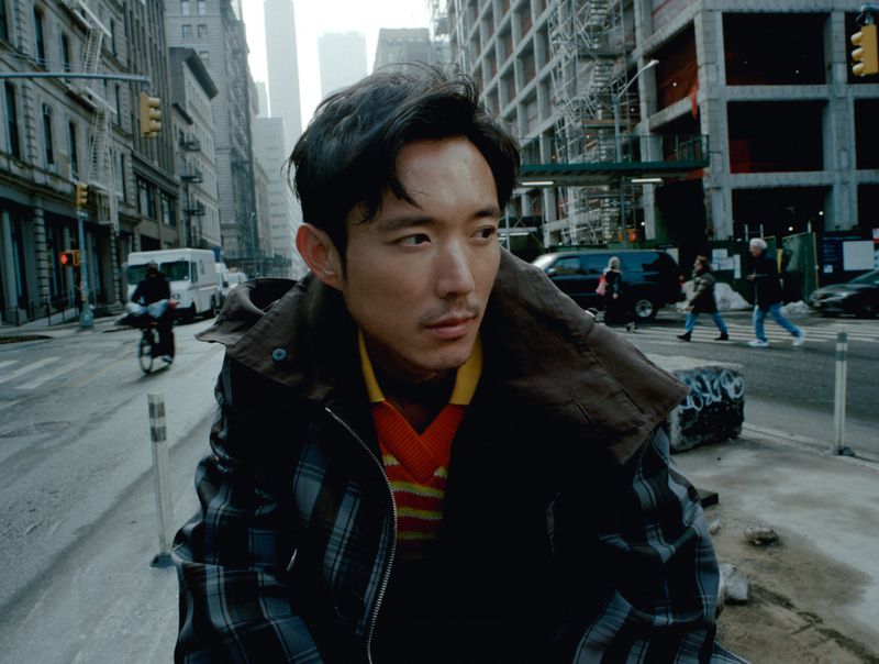 Portrait of Asian man in plaid coat standing on city street with high-rise buildings and pedestrian crossing in the background.