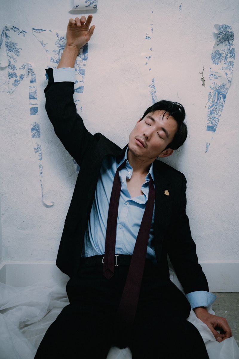 Portrait of a dark-haired man in a black suit lying next to a wall with peeling wallpaper and a white sheet on the floor.