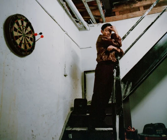 Actor Justin H. Min standing on an indoor staircase beneath a dartboard for Cero Magazine, photographed by Elinor Kry.