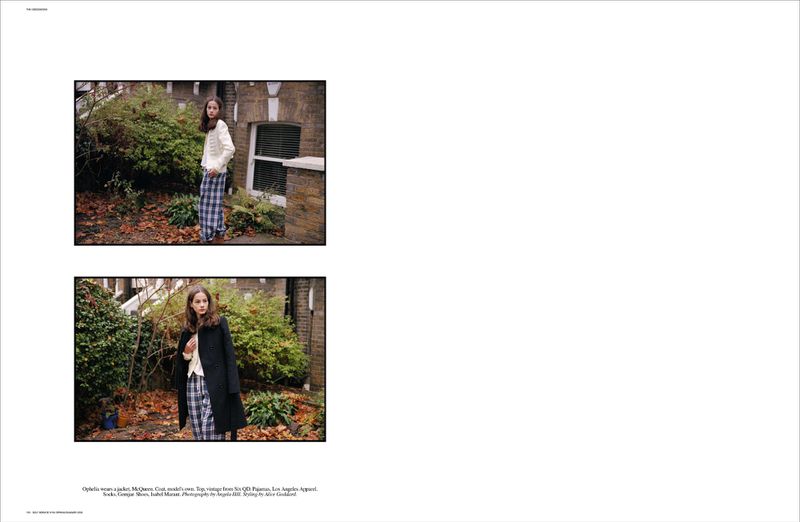 Young woman in fashionable, relaxed attire posing outdoors on a stone path near foliage and a brick building in daytime lighting.
