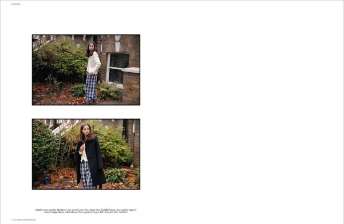 Young woman in fashionable, relaxed attire posing outdoors on a stone path near foliage and a brick building in daytime lighting.