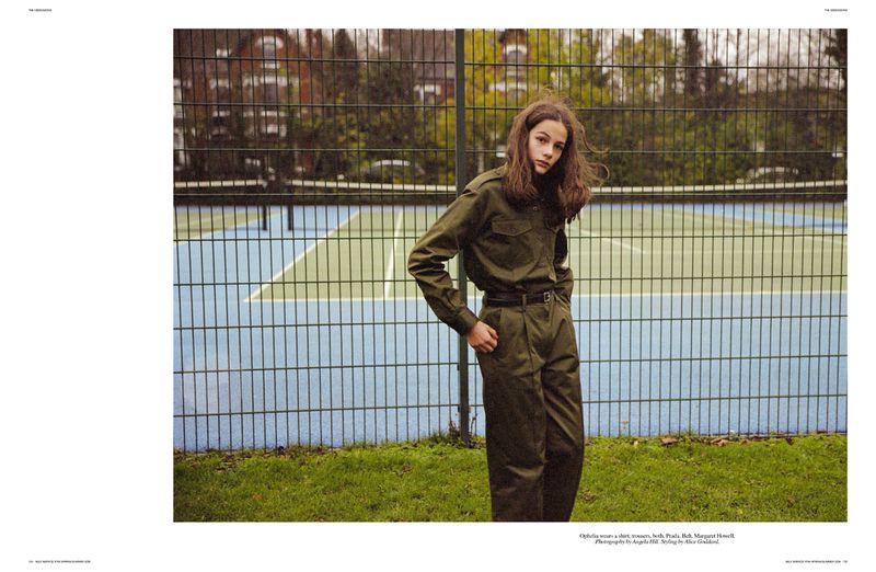 A teenage woman in a dark olive green jumpsuit stands on grass in front of a blue and green tennis court fence.