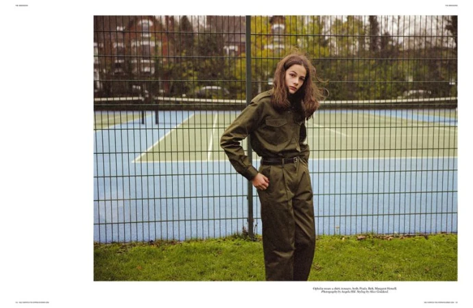 A teenage woman in a dark olive green jumpsuit stands on grass in front of a blue and green tennis court fence.