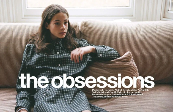 Young woman with long brown hair sits on a beige corduroy couch wearing a green and black plaid shirt dress in an indoor setting.
