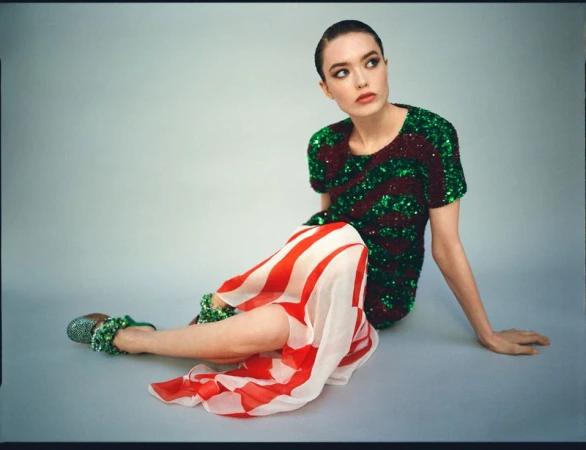 A stylish woman with slicked-back hair poses in a studio wearing a green and red sequined top and a red and white striped skirt.