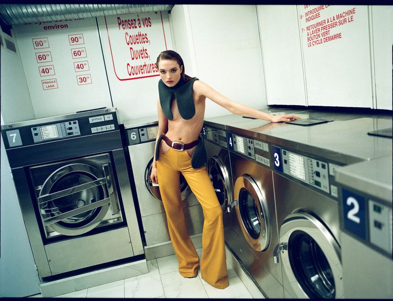A woman in yellow pants poses in a laundromat by a row of stainless steel washing machines, with vintage color grading and fashion style.