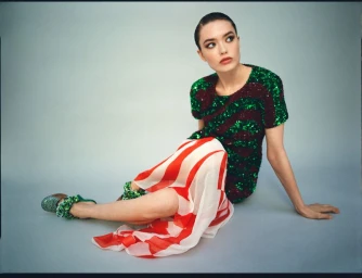 Model seated on a grey floor wearing a sequined green and burgundy top with a red and white striped skirt against a plain studio background.