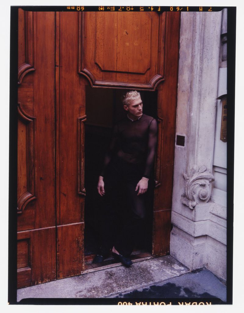 A person stands in an open wood door wearing a black mesh top and pants, posing for a fashion editorial shot outdoors.