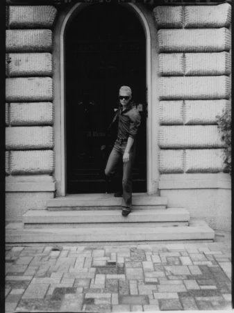 Adult male in jeans, shirt, sunglasses walking out of an archway doorway in an outdoor setting with brick and stone structures.