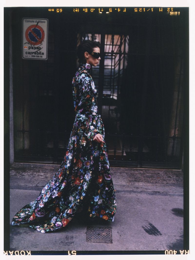A woman in a floral dress walks in front of a dark building in a daytime urban setting. She wears sunglasses and has her hair up.