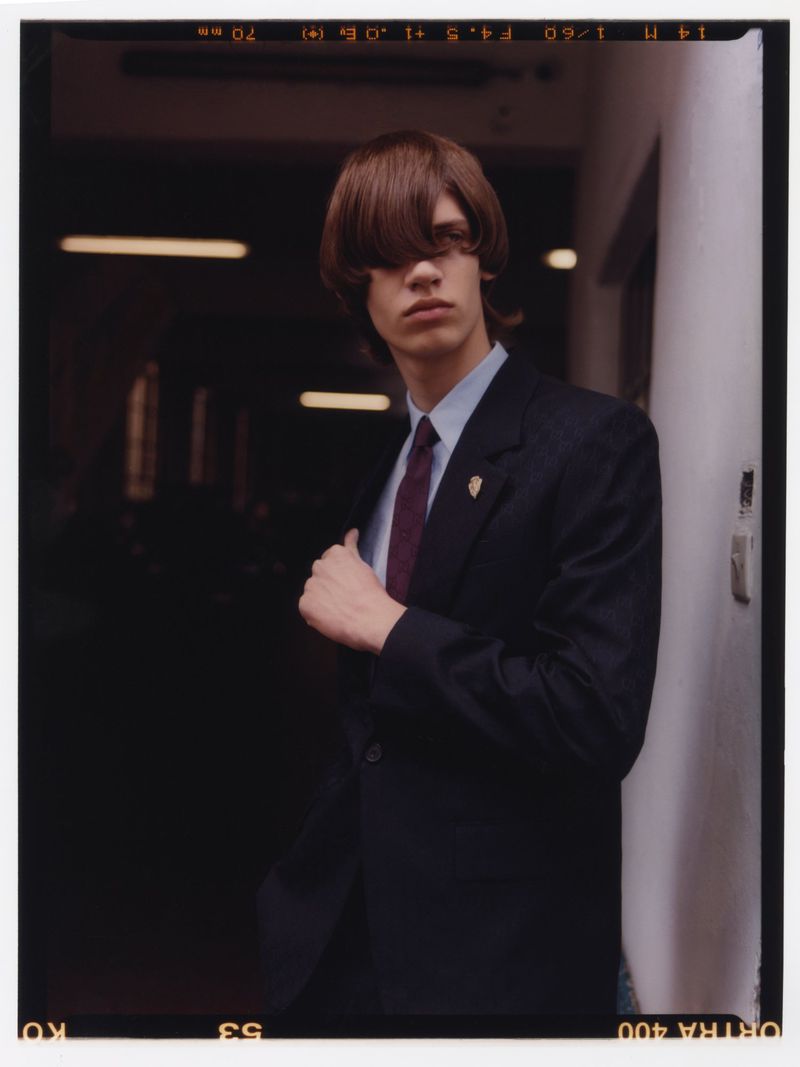Portrait of a young adult with long brown hair wearing a dark suit, light blue shirt, and tie, posed in a dimly lit hallway.