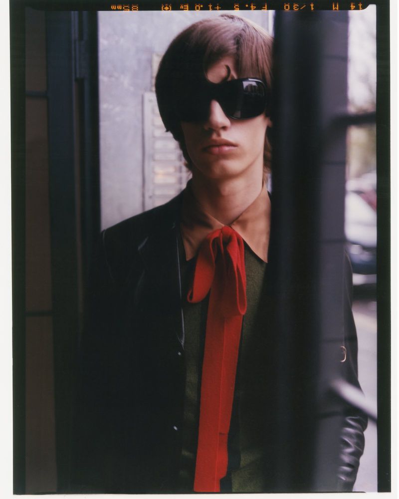 A young adult with dark hair and sunglasses poses in a suit with a long red tie in front of a textured wall with metal accents.