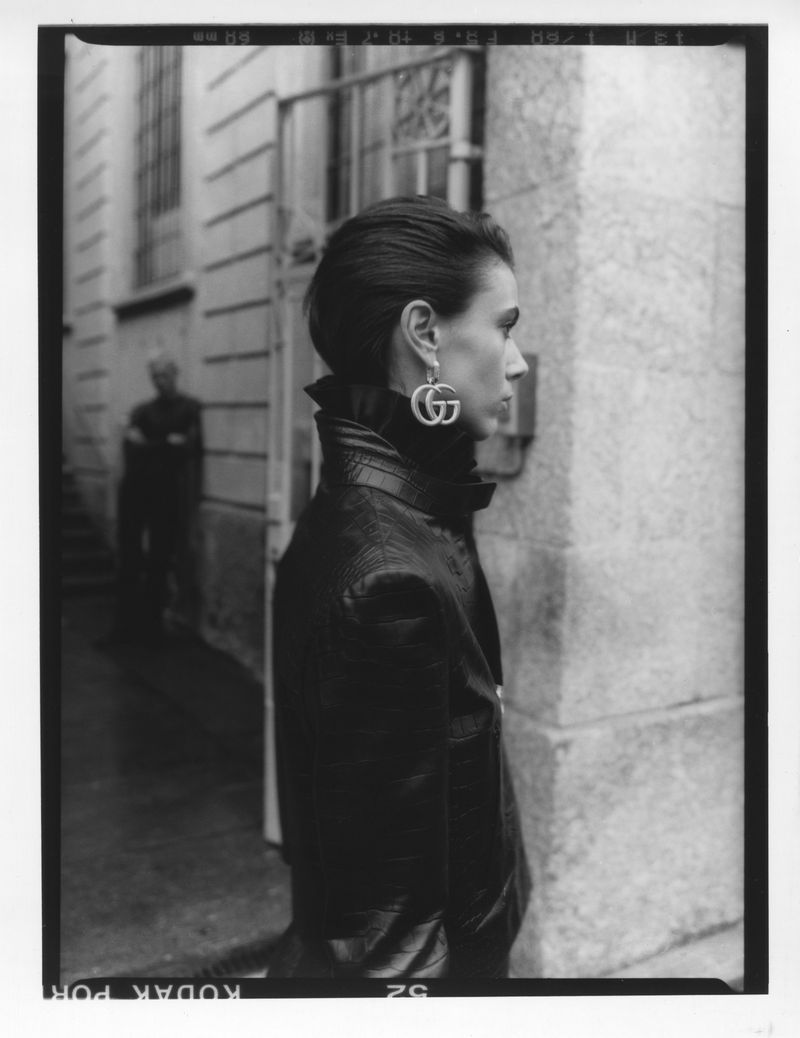 Black and white portrait of a woman wearing statement logo earrings and a dark jacket against a stone building exterior.