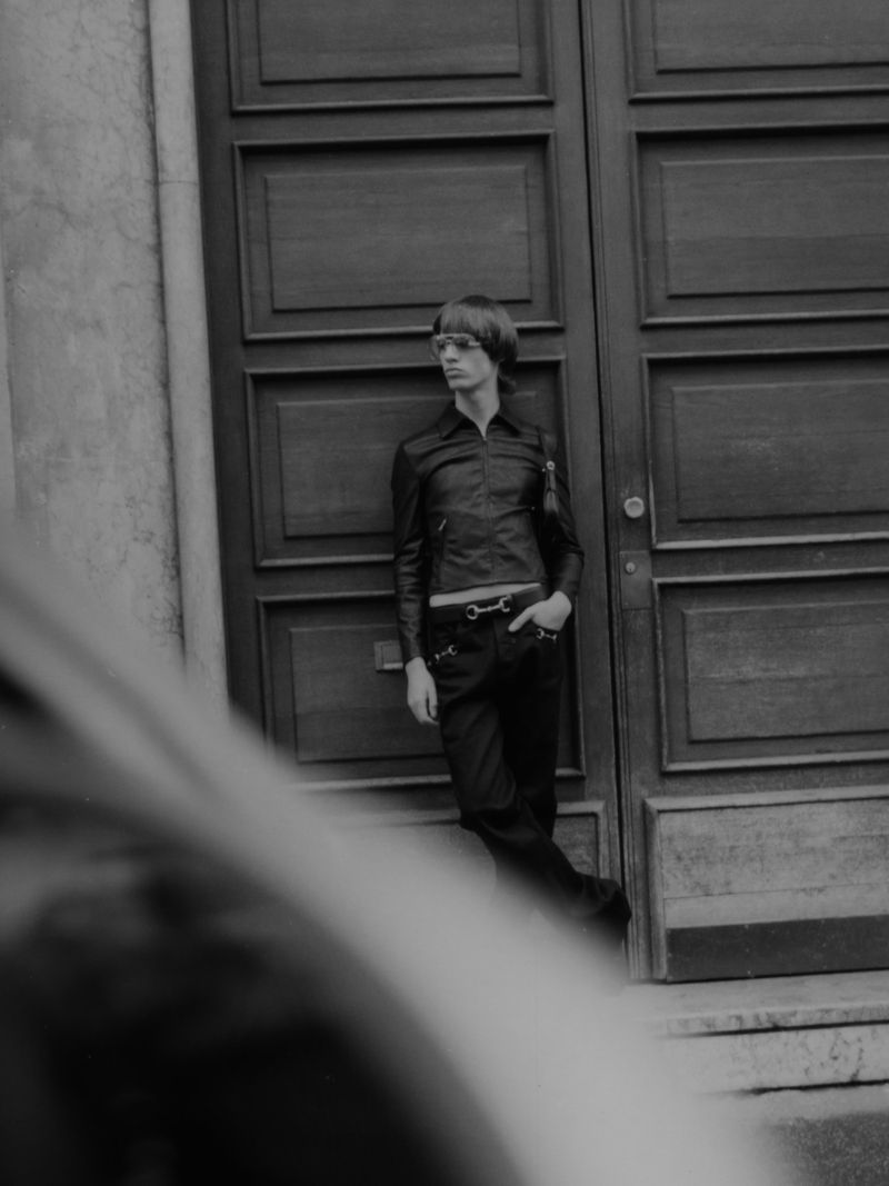 A monochromatic shot shows a slim adult man in a leather jacket leaning against a panelled door in an urban setting.