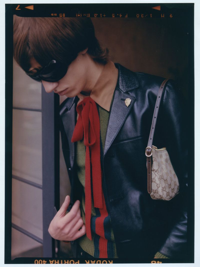 Stylish adult with a red scarf, wearing sunglasses and a black jacket, carrying a Gucci handbag in an indoor setting with soft lighting.