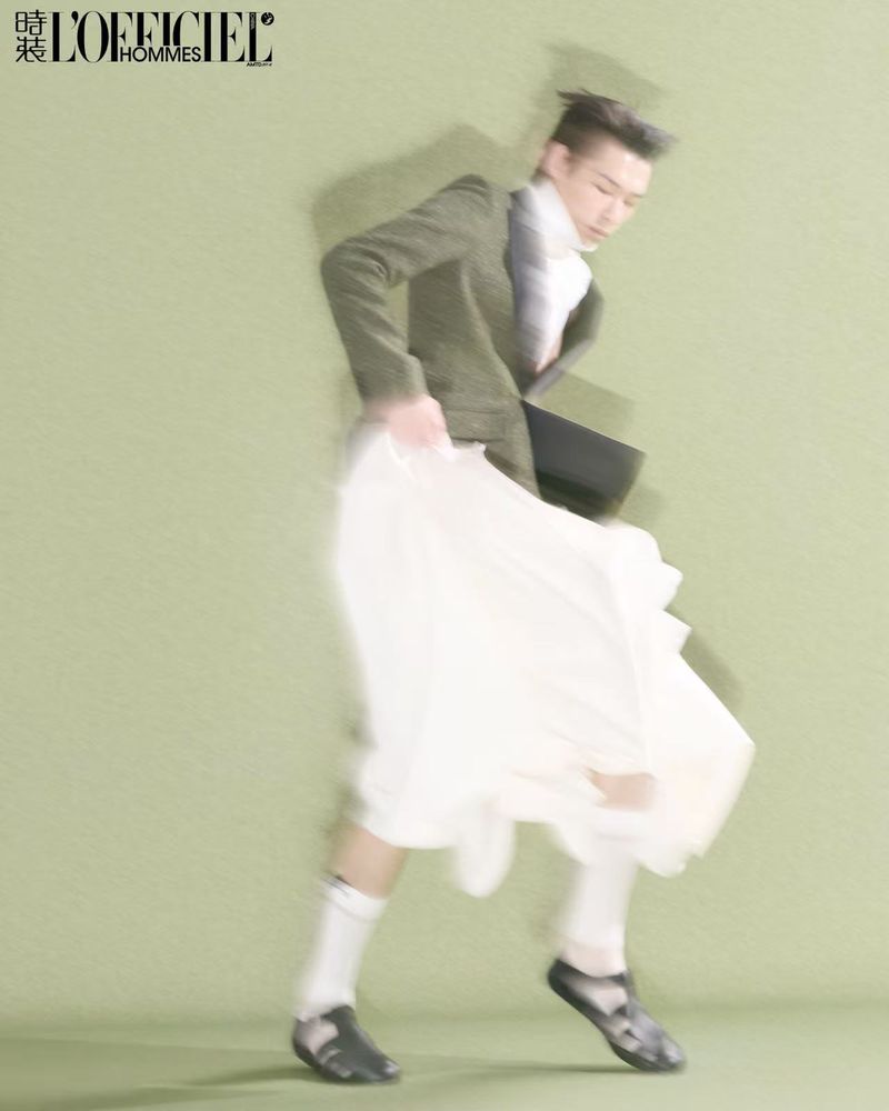 A person in a motion-blurred green blazer and white skirt dancing against a solid light green background.
