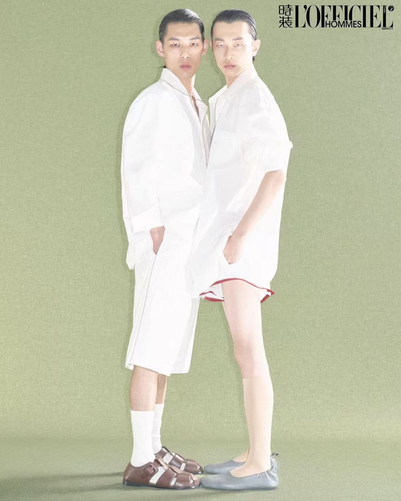 Two androgynous Asian men in white shirts and shorts pose against a green background, showcasing modern fashion and minimalist styling.