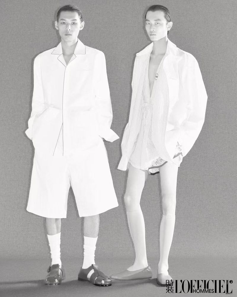 Two adult Asian men pose in white fashion against a gray background, wearing shorts, open shirts, socks, and casual shoes in a studio shot.