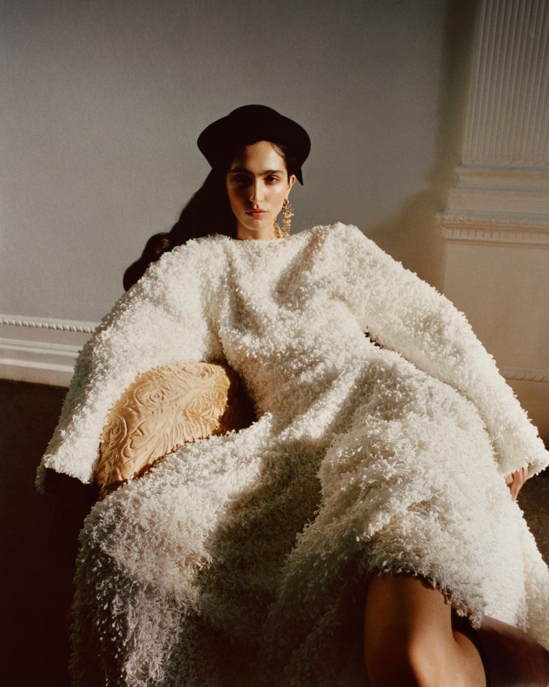 A woman wearing a textured white fringe dress and dark beret sits in an armchair against a plain wall, captured in soft golden light.