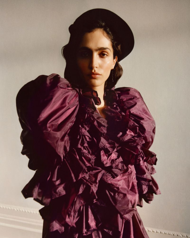 A woman with dark hair wearing a black hat and a plum-colored, ruffled dress poses in a studio with natural side lighting.