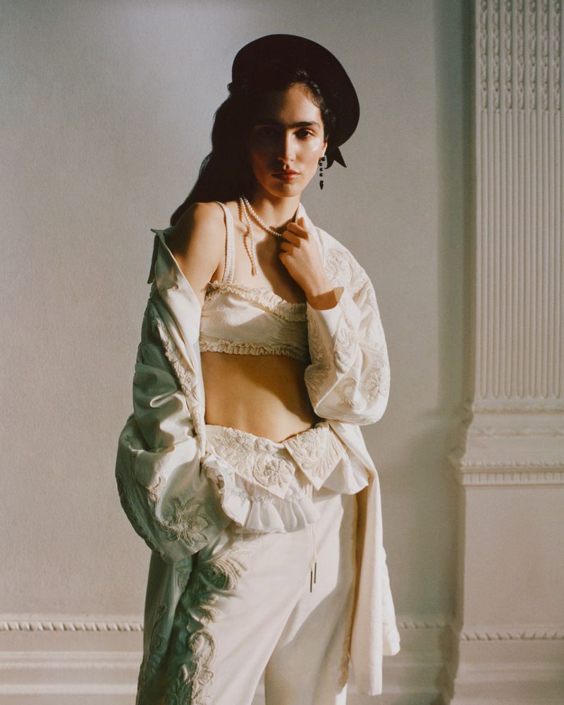 Woman in ornate white outfit poses indoors with black hat, jewelry, and soft light on architectural background. Fashion editorial shot.
