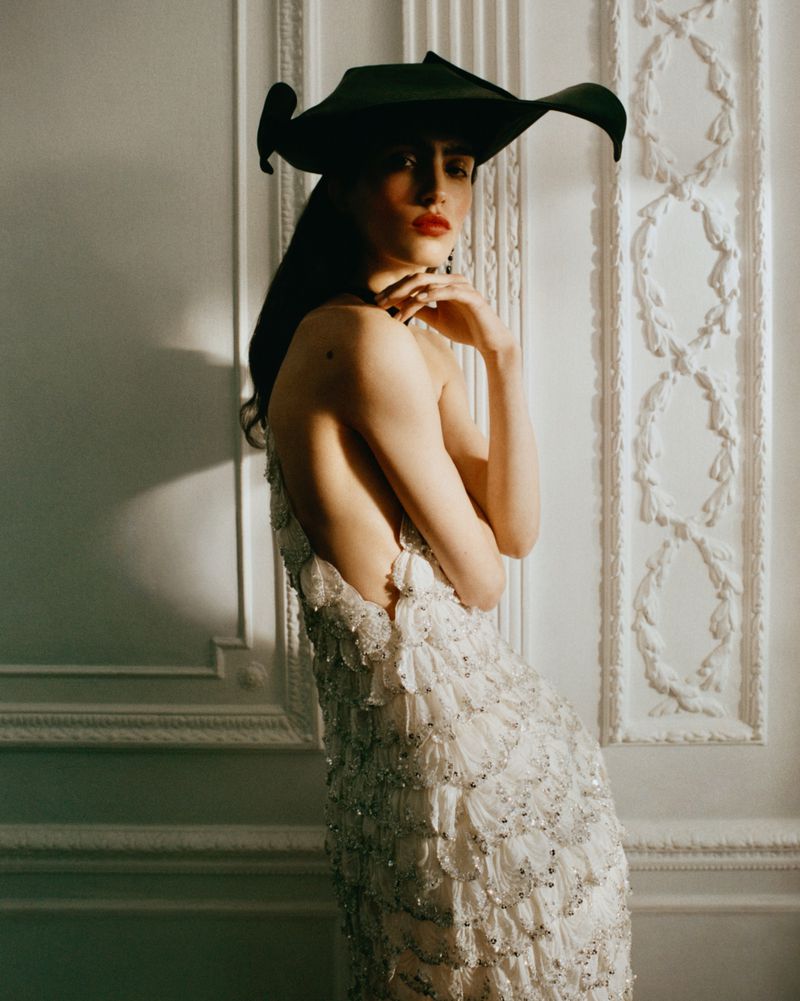 A fashionable woman with red lipstick, wearing an ornate white dress and wide-brimmed black hat, poses in front of a patterned white wall.
