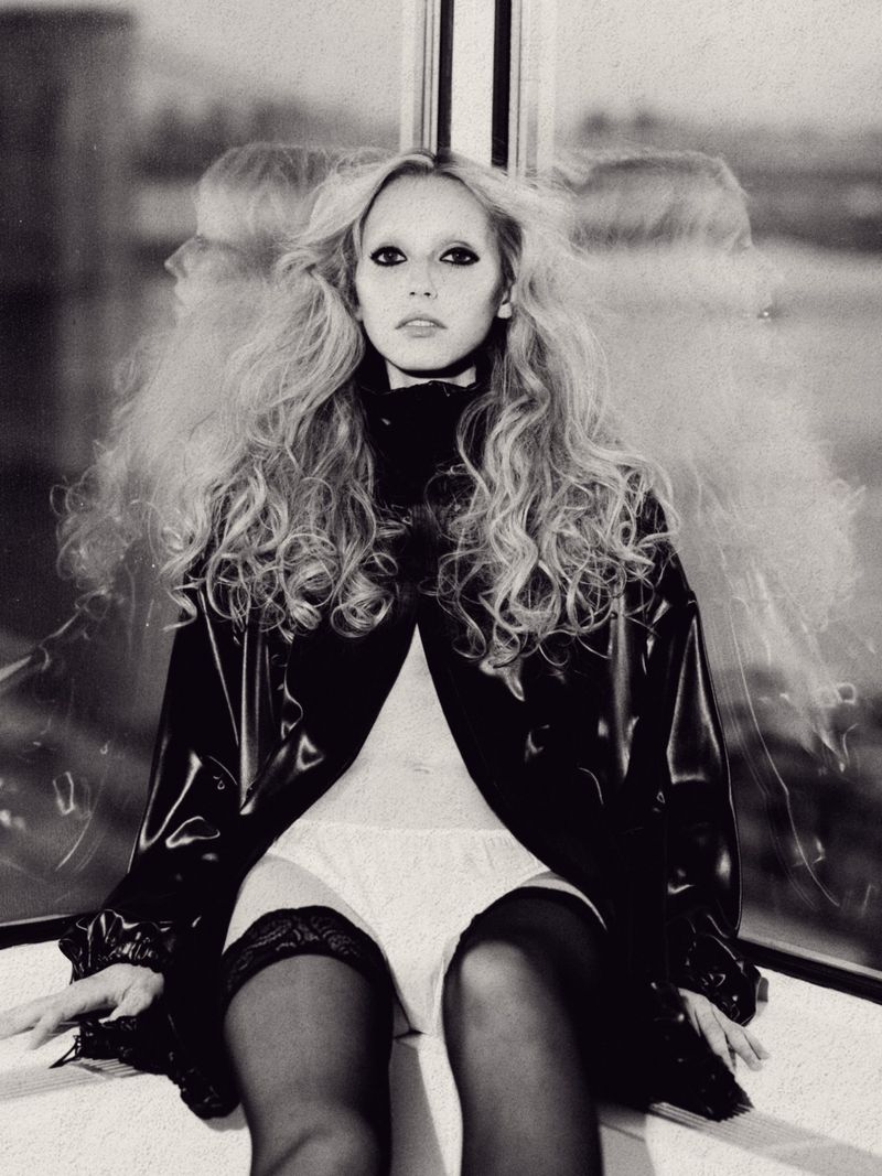 Monochrome studio portrait of a young adult woman with long wavy blonde hair, wearing a shiny black jacket and thigh-high stockings.