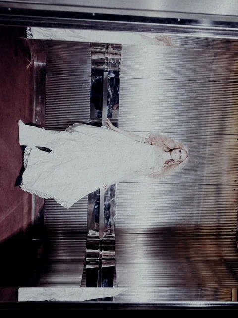 Fashion editorial shot of a slender woman in a white lace dress lying in a silver elevator with red flooring, captured with soft lighting.