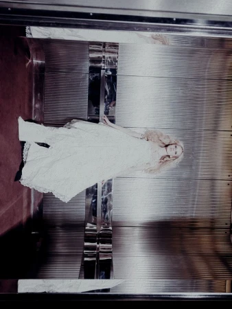 Fashion editorial shot of a slender woman in a white lace dress lying in a silver elevator with red flooring, captured with soft lighting.