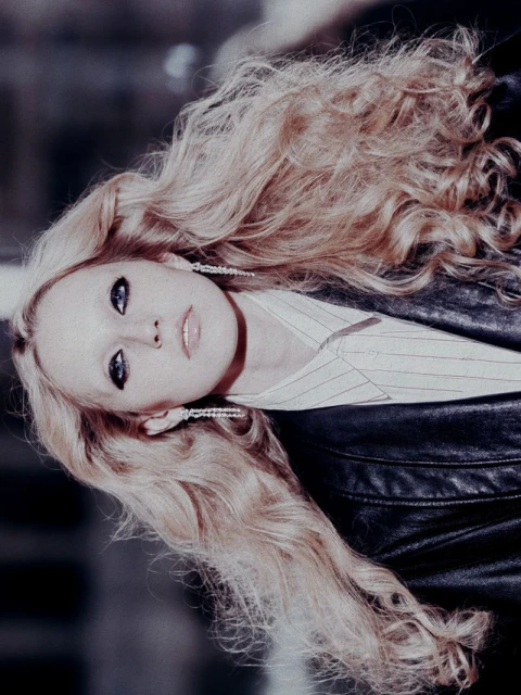 Portrait of a blonde woman with long wavy hair, wearing a black leather jacket and striped shirt, gazing up at the camera.