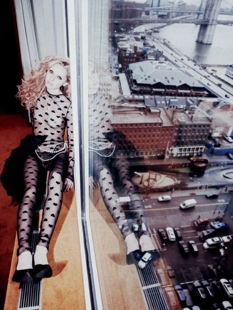 Blonde fashion model in patterned top and leggings poses next to a window with city view and reflection, casting a moody shadow.