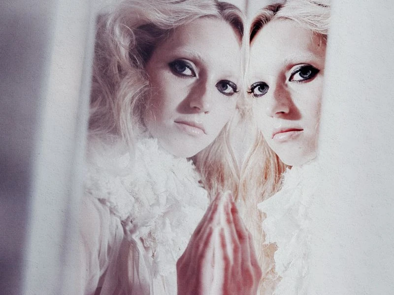 Close-up portrait of a fair-skinned woman with heavy eye makeup reflected in a mirror against a white backdrop.