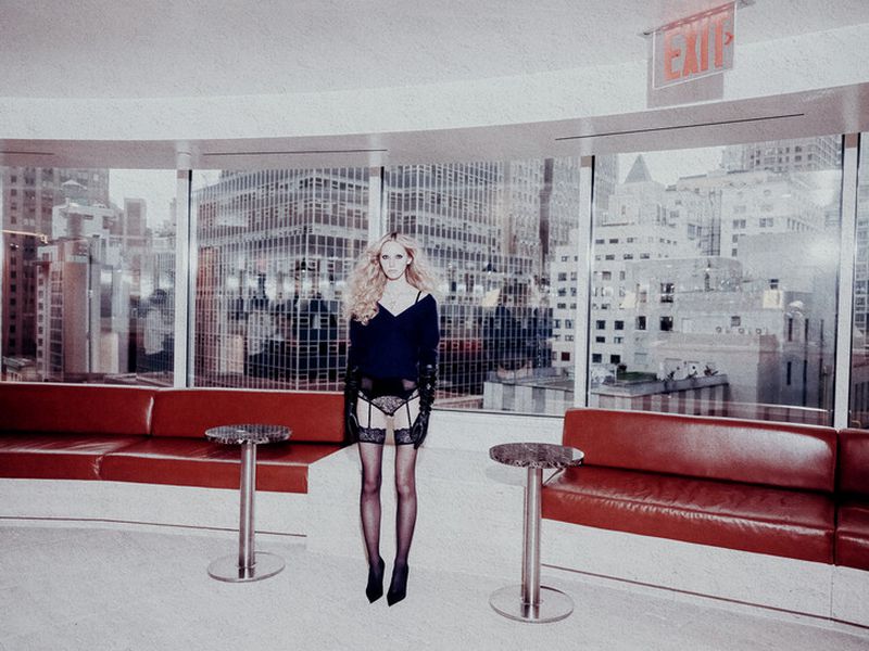 Woman in lingerie poses in an interior space with large windows overlooking a city skyline; red sofas flank, and the light is soft.