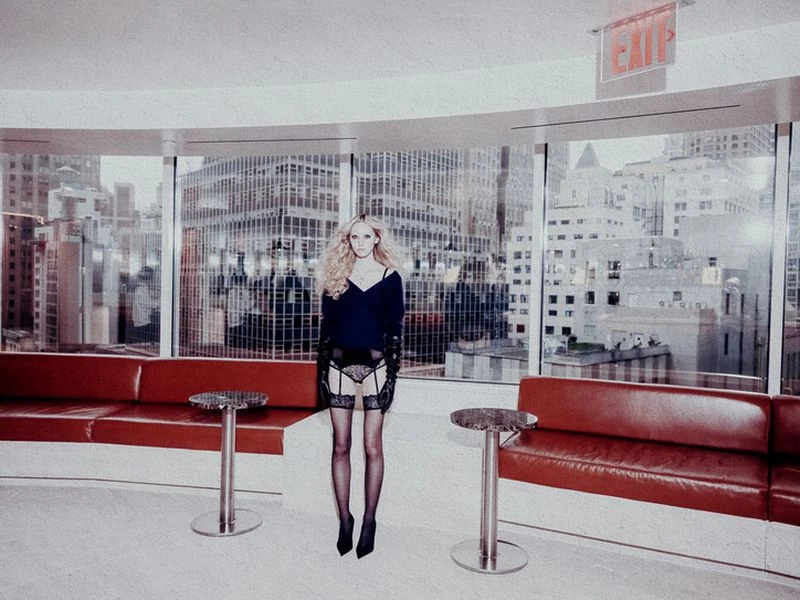 Woman in lingerie poses in an interior space with large windows overlooking a city skyline; red sofas flank, and the light is soft.