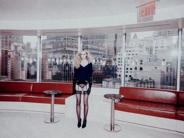 Woman in lingerie poses in an interior space with large windows overlooking a city skyline; red sofas flank, and the light is soft.