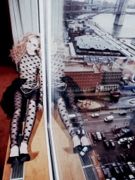 Grace Van Petten wearing a sheer heart print bodysuit seated by a large window overlooking an urban street view.
