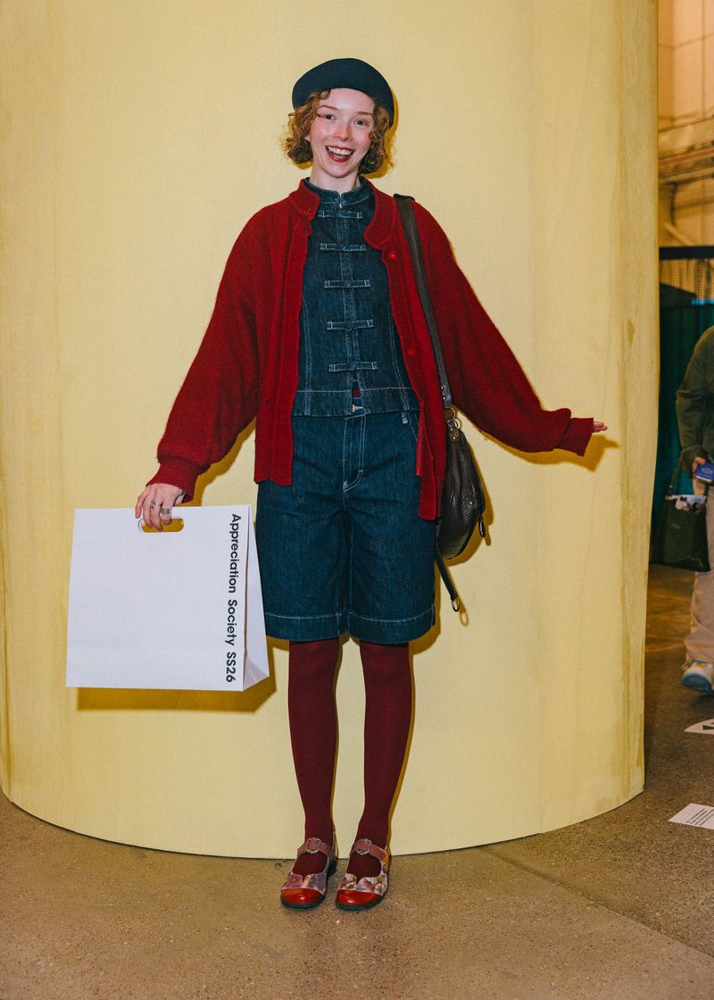 A woman in a red cardigan and denim jumpsuit stands in front of a yellow wall, holding a white shopping bag and a brown purse.