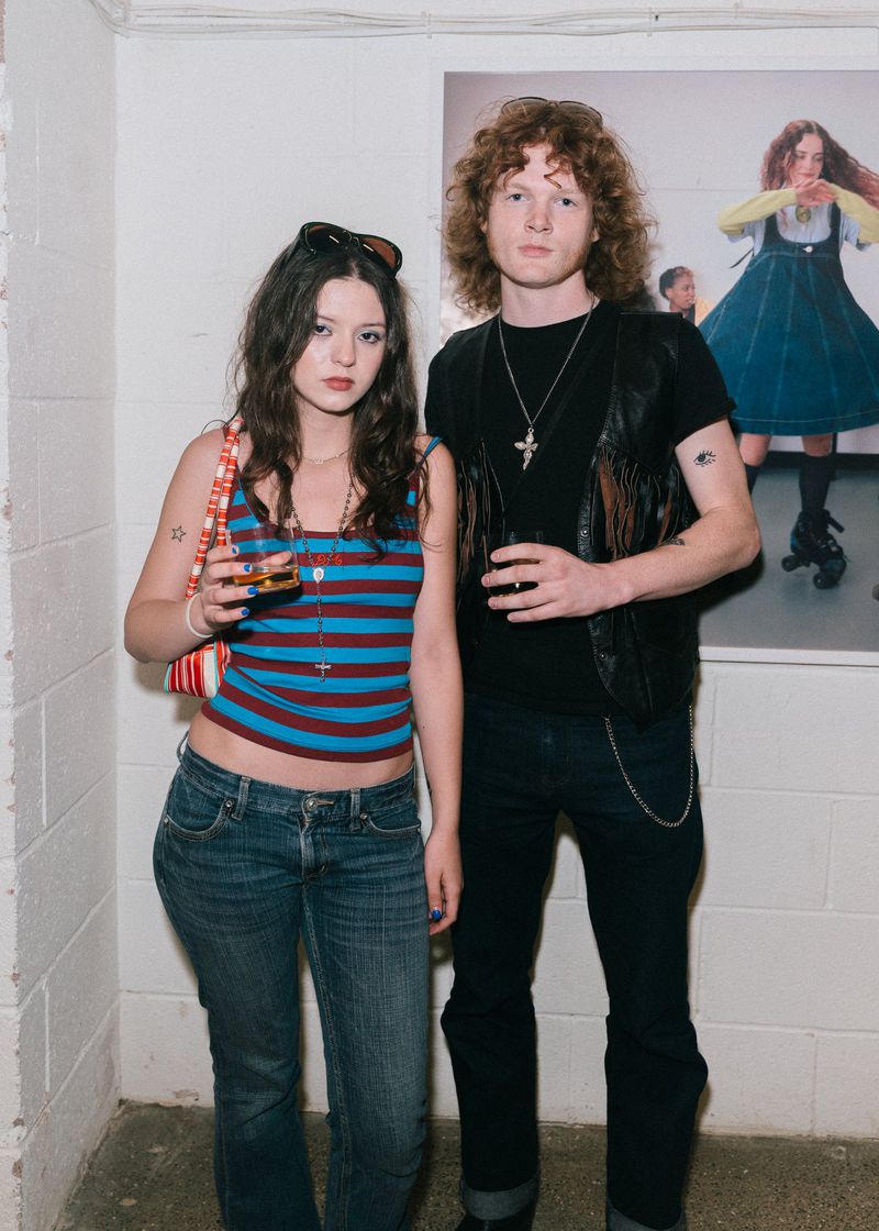 Young woman and man pose indoors, she wears a striped top and jeans, he a vest and dark pants, with an art piece in the background.