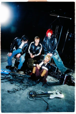 Band of four young men wearing leather jackets in a music studio with instruments and cables, posing for a photo.