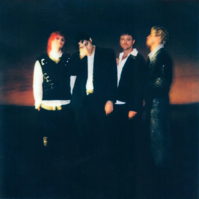 Four people in a dark indoor space. Three are wearing blazers, with a fourth person wearing a vest and white shirt.