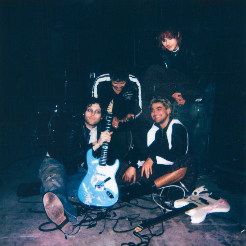 Four people posing indoors in a dark room with music equipment including a guitar, drums, and cables. Band promo shot.