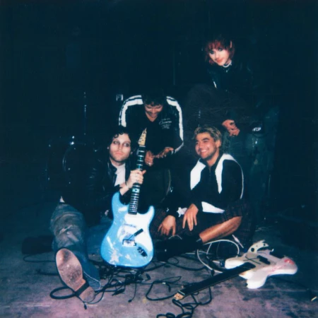 Four people posing indoors in a dark room with music equipment including a guitar, drums, and cables. Band promo shot.