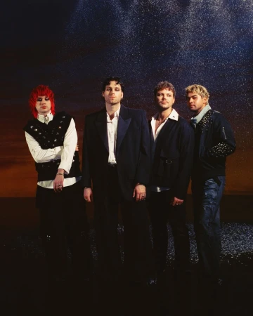 A group of four adult male musicians stand indoors in formal attire and fashionable hairstyles against a starry backdrop.