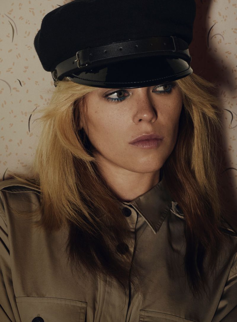 Close-up portrait of a woman with blonde hair wearing a black cap and a tan-colored shirt. She is photographed indoors against a patterned wallpaper.