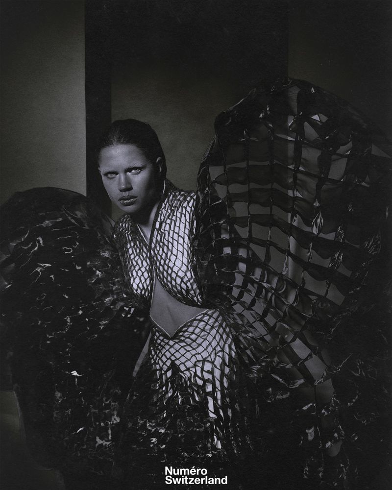 Monochrome portrait of an adult woman in a patterned bodysuit, framed by large abstract sculptural pieces. Fashion photography.