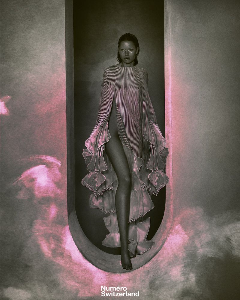 Fashion shot of a model in a pleated pink gown within an architectural oval alcove, striking a pose under studio lighting for Numero Switzerland.