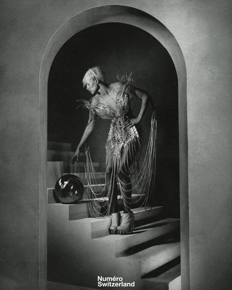 Monochrome fashion shot of an adult in a decorative dress and platform boots, touching a silver sphere on stairs beneath an archway.