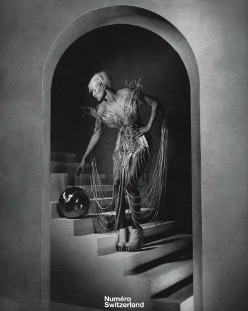 Monochrome fashion shot of an adult in a decorative dress and platform boots, touching a silver sphere on stairs beneath an archway.