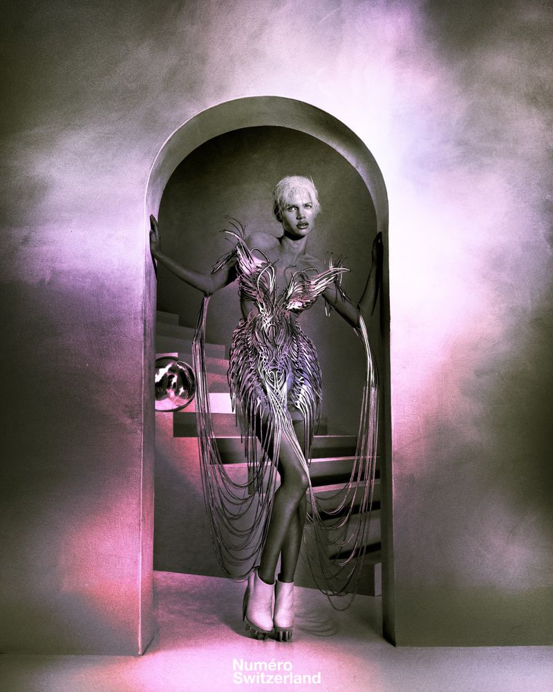 A slender woman in a futuristic silver and white dress with trailing strands stands framed by an arched doorway with silver stairs in the background.