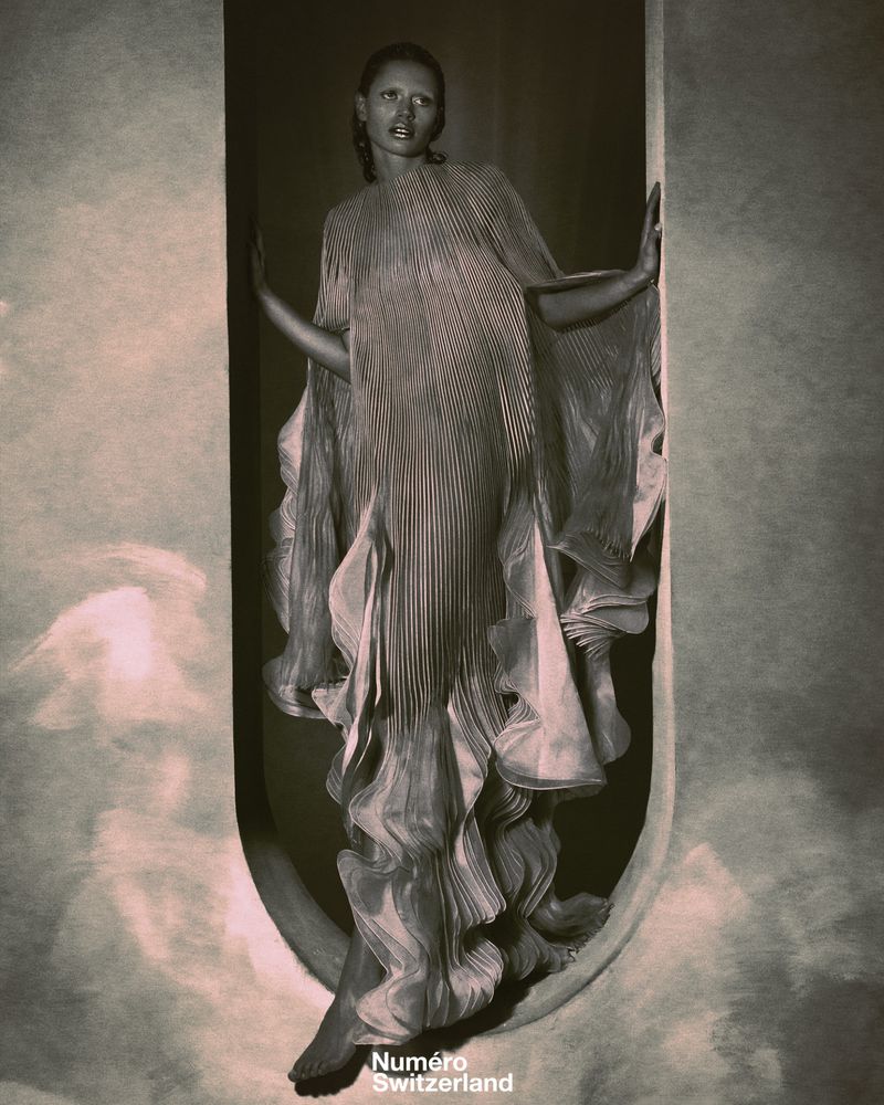 Fashion editorial shot of a Black female model posing in a pleated gown within a curved architectural frame, in a dramatic, grey-toned indoor setting.